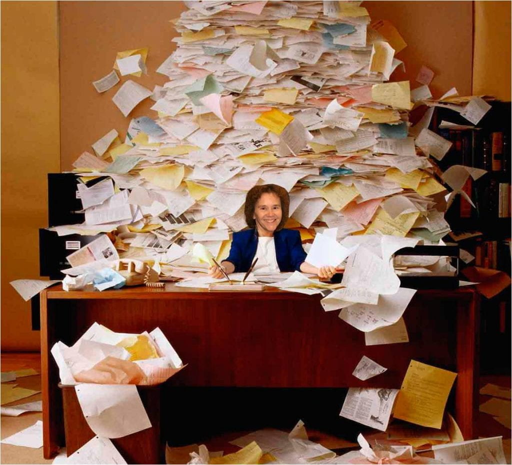 Person sitting behind a desk with a mountain of paperwork.